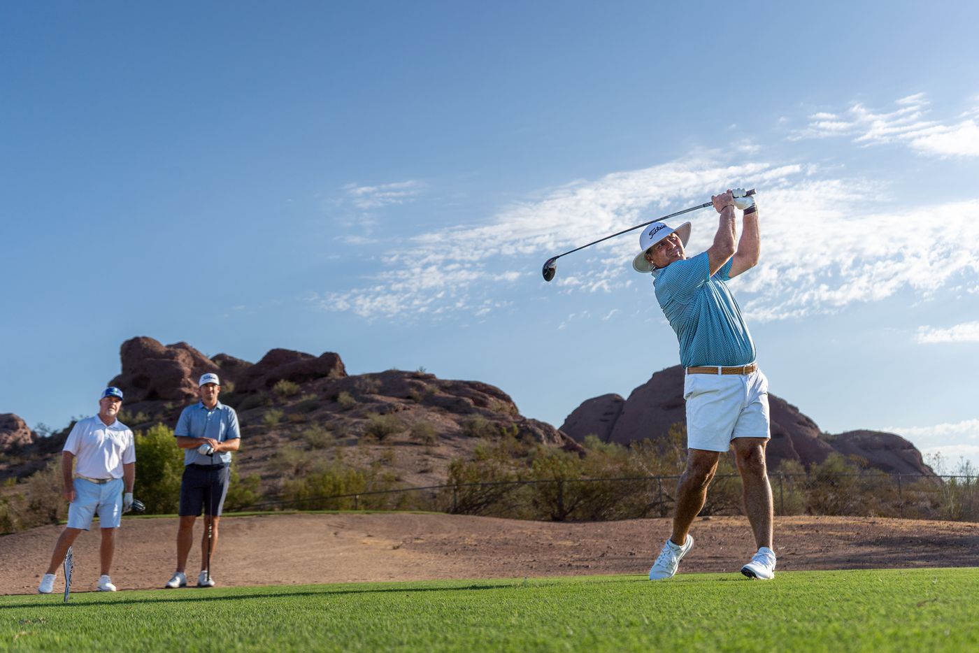 Golfer teeing off in front of red desert buttes as his group looks on